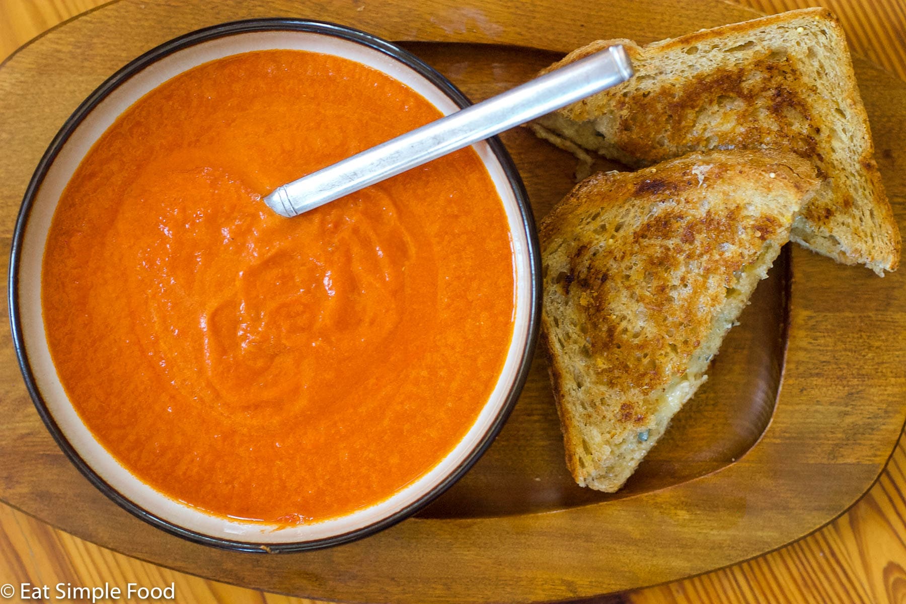 White bowl with black rim of red tomato bisque soup on a wood rectangle plate. Grilled cheese sandwich cut in half diagonally on the side. Top view.