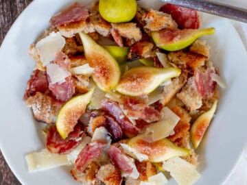 Top view of a white plate with fork with quartered figs, sliced salami, shaved parmesan and croutons.