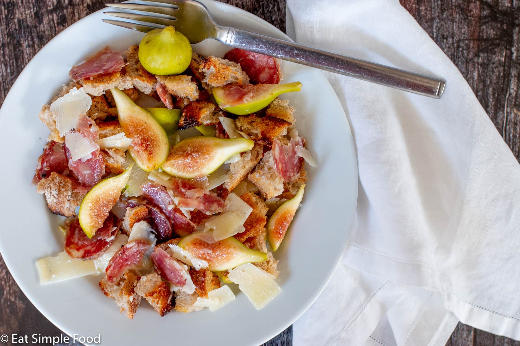 Top view of a white plate with fork with quartered figs, sliced salami, shaved parmesan and croutons.