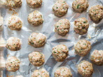Sheet pan lined with aluminum foil with cooked meatballs with basil on it. Top view.