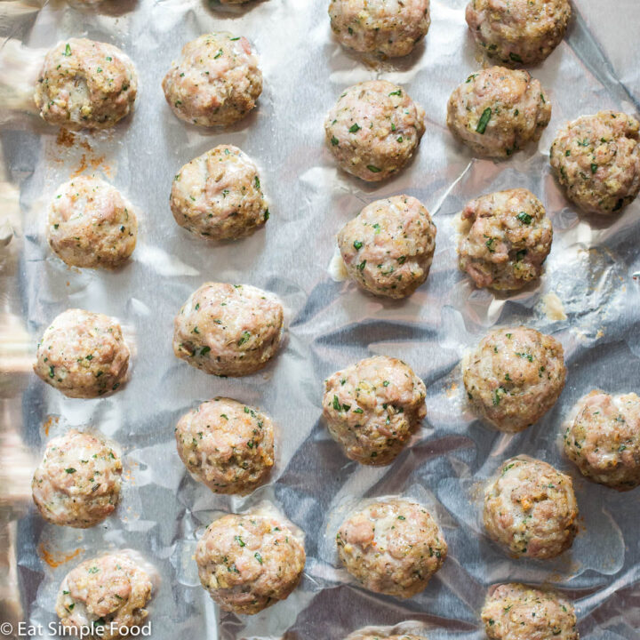 Sheet pan lined with aluminum foil with cooked meatballs with basil on it. Top view.