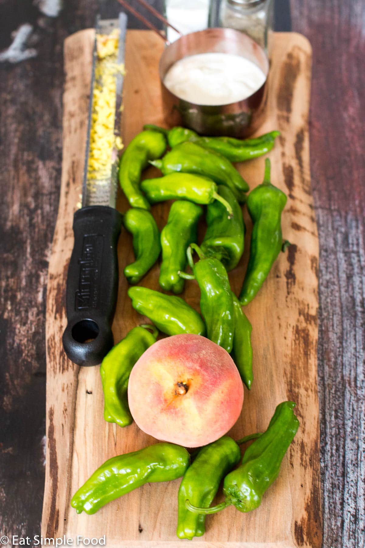 Live edge wood cutting board with 12 small green peppers, one peach, ½ cup of white yogurt, and a microplane zester with grated ginger.