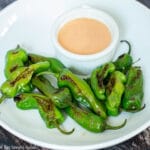 White plate of brown seared / blistered small green peppers with a small white bowl of light orange peach sauce.