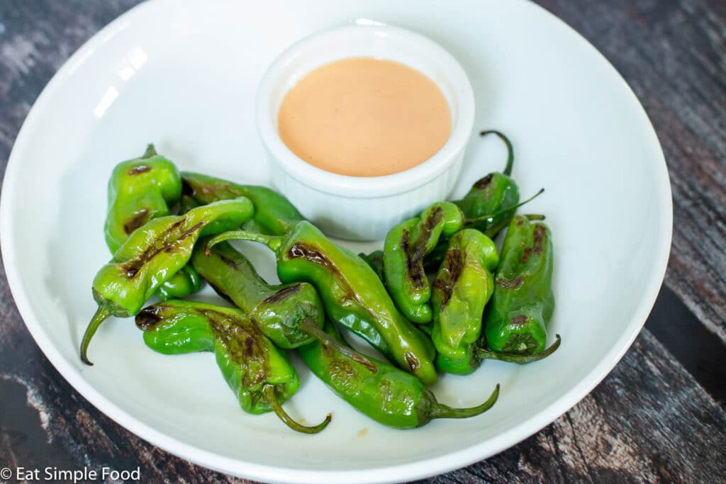 White plate of brown seared / blistered small green peppers with a small white bowl of light orange peach sauce.