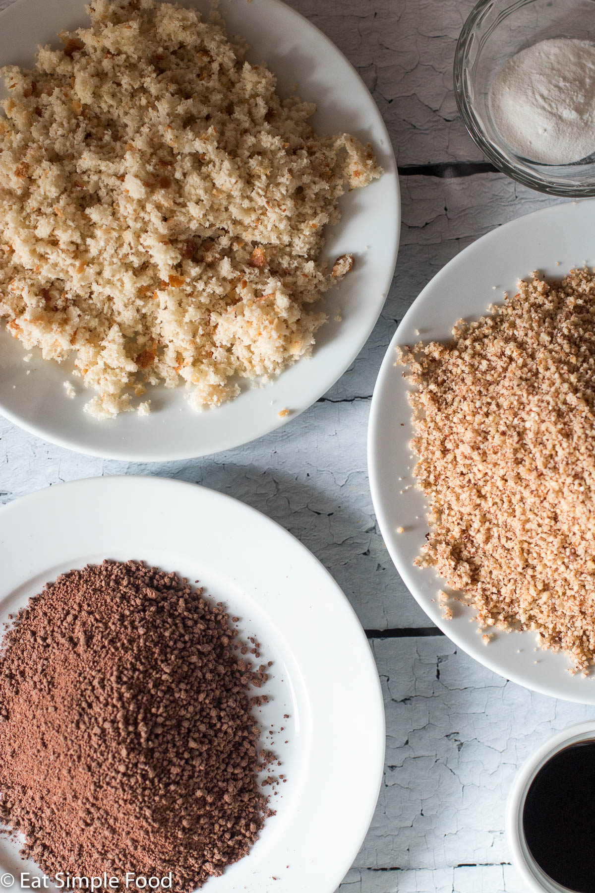 Ground up cake ingredients on white plates: breadcrumbs, hazelnuts, chocolate chips. All pulverized.