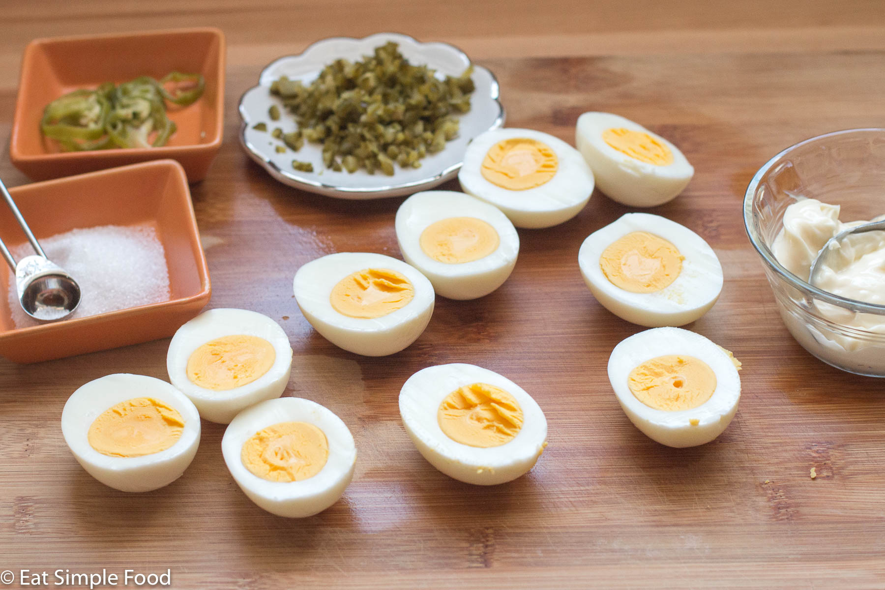 10 boiled eggs peeled and halved on a wood cutting board with a small bowl of mayonnaise, a container of salt with ¼ tsp, sliced peppers, and chopped pickles.