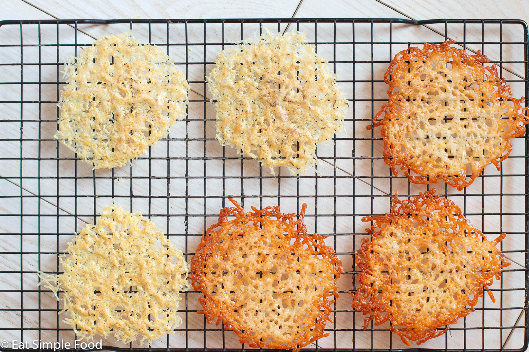 6 Parmesan crisps on an elevated wire rack on a white table. 3 are darker and 3 are lighter in color.