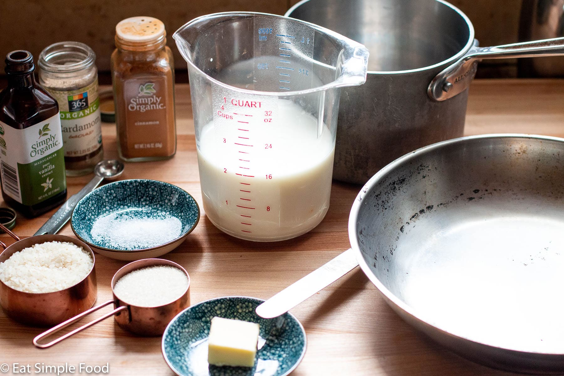 Raw ingredients in small containers, measuring cups, and 1 pot and 1 pan. In containers are sugar, milk, butter with knife and salt.