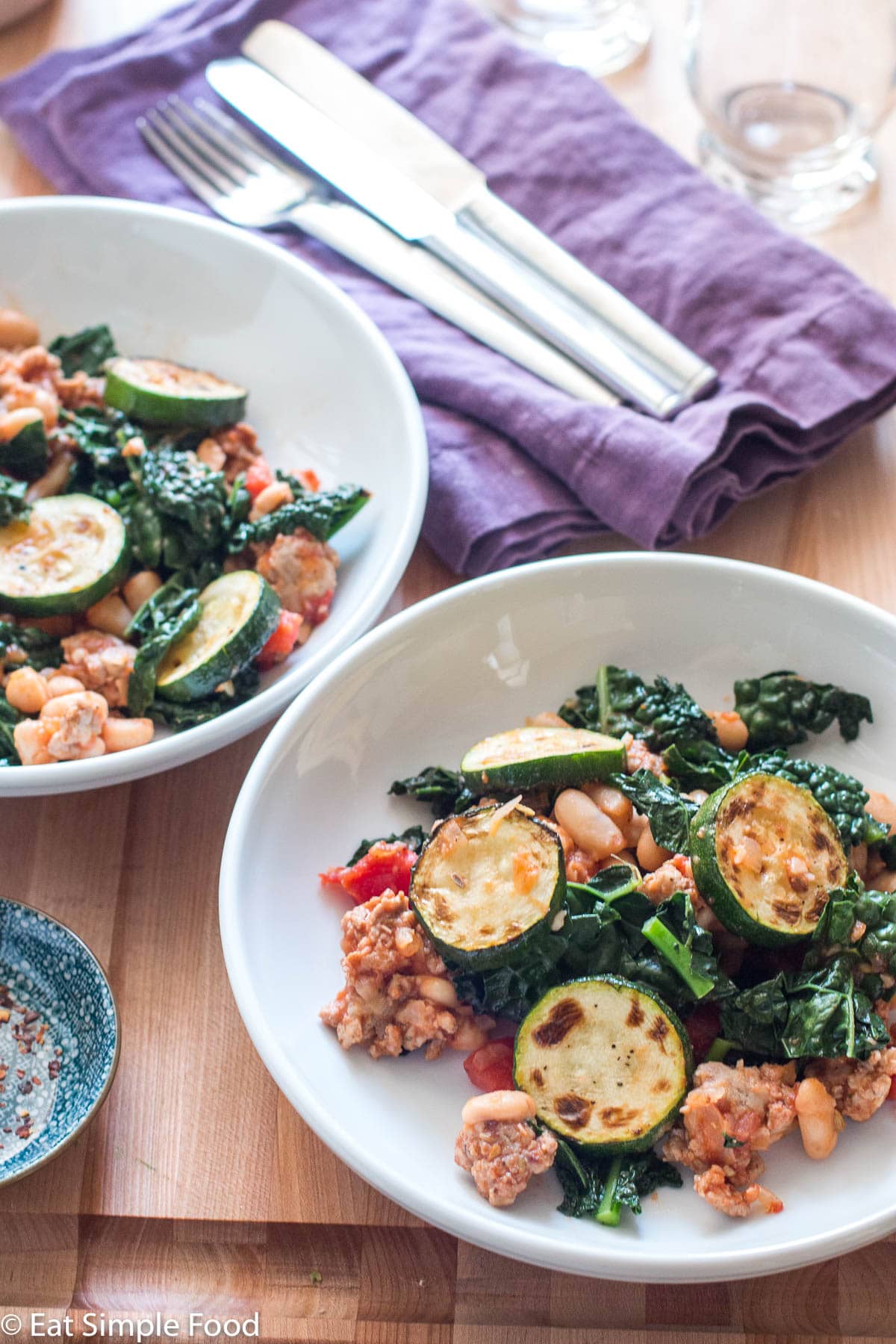 Side view of two plates of ragout: cooked sausage, sliced seared zucchini, chopped kale, white beans, and tomato. Side of hot chili flakes on the side. Napkins, silverware, and glasses in background.