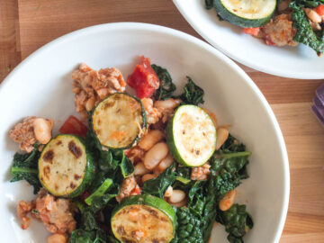 Top down view of two plates of ragout: cooked sausage, sliced seared zucchini, chopped kale, white beans, and tomato. Side of hot chili flakes on the side.