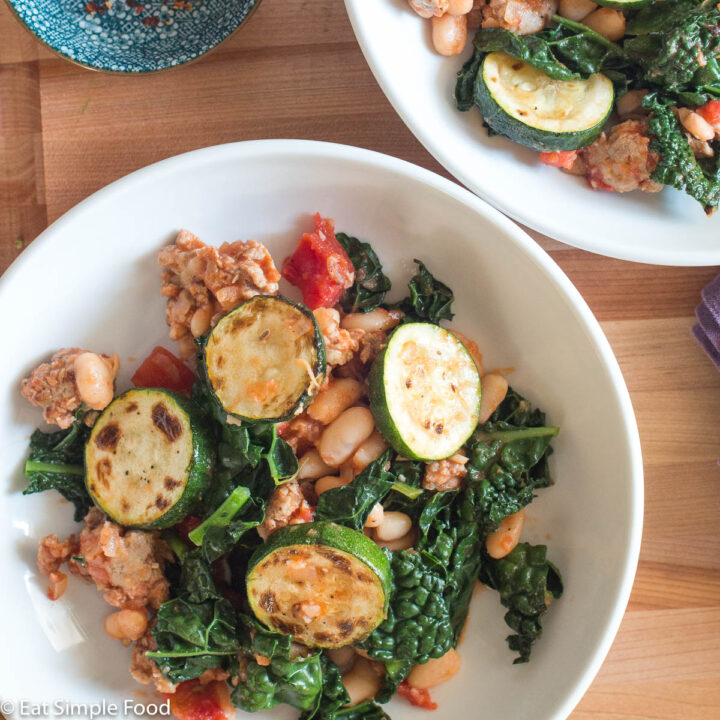 Top down view of two plates of ragout: cooked sausage, sliced seared zucchini, chopped kale, white beans, and tomato. Side of hot chili flakes on the side.