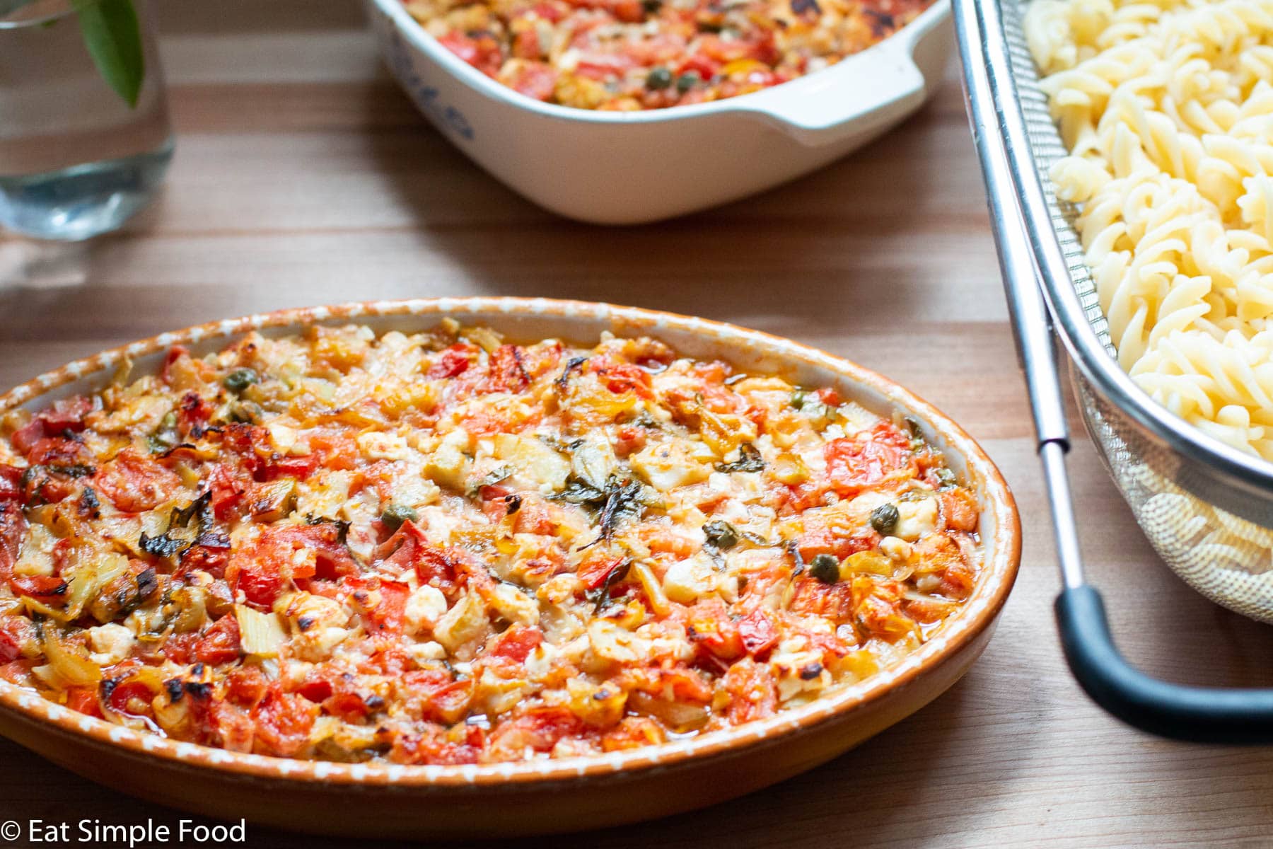 2 baking casseroles filled with cooked diced tomato, artichoke heart, feta, capers, basil, and peppers. Browned on top. Basil and fusilli pasta on side.