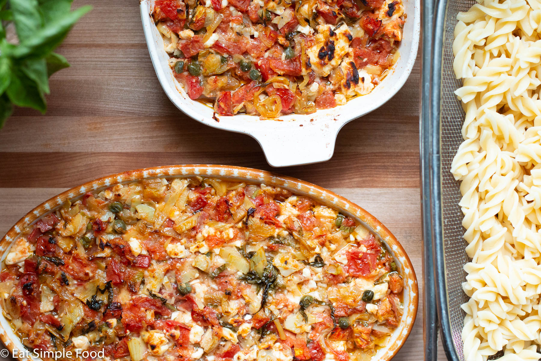2 baking casseroles filled with cooked diced tomato, artichoke heart, feta, capers, basil, and peppers. Browned on top. Basil and fusilli pasta on side.