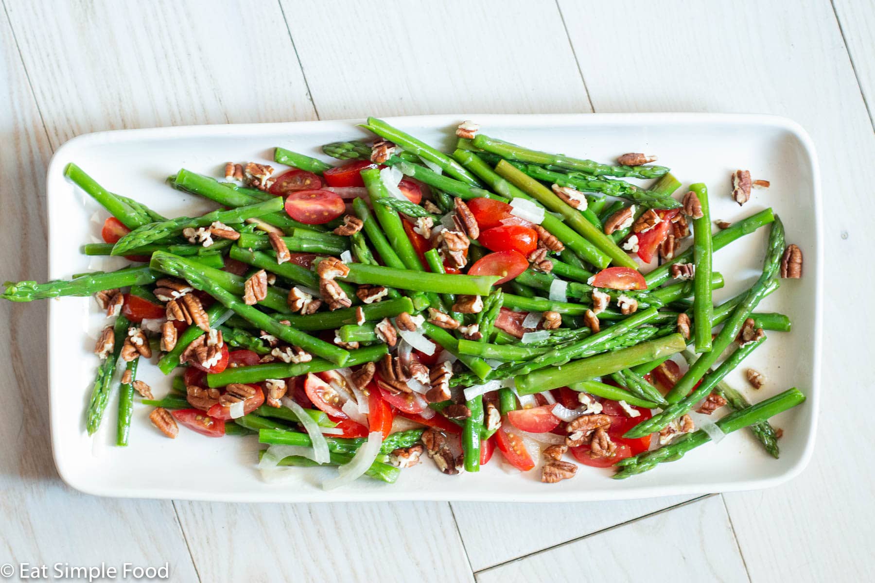 Top view of long white plate with blanched asparagus, raw halved cherry tomatoes, sliced onions, and chopped pecans.