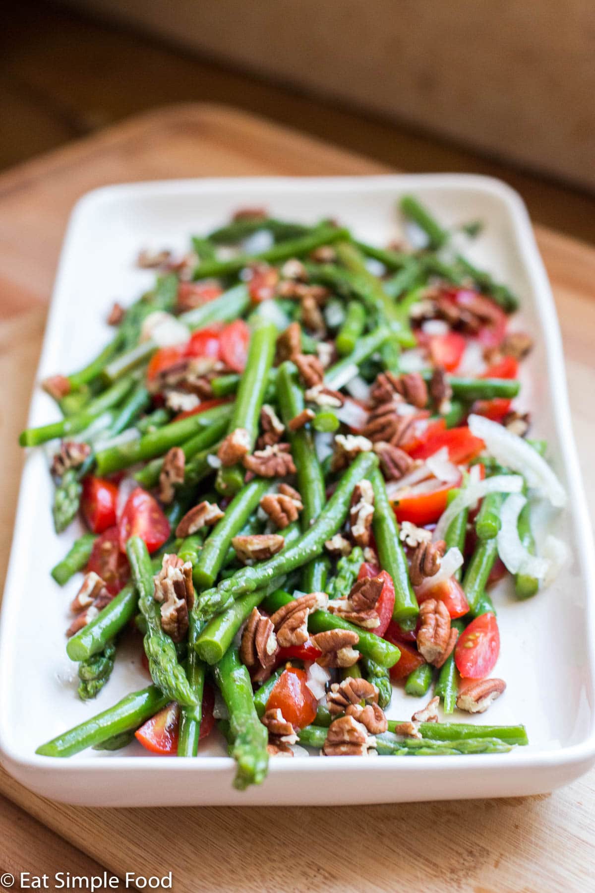 Side view of long white plate with blanched asparagus, raw halved cherry tomatoes, sliced onions, and chopped pecans.