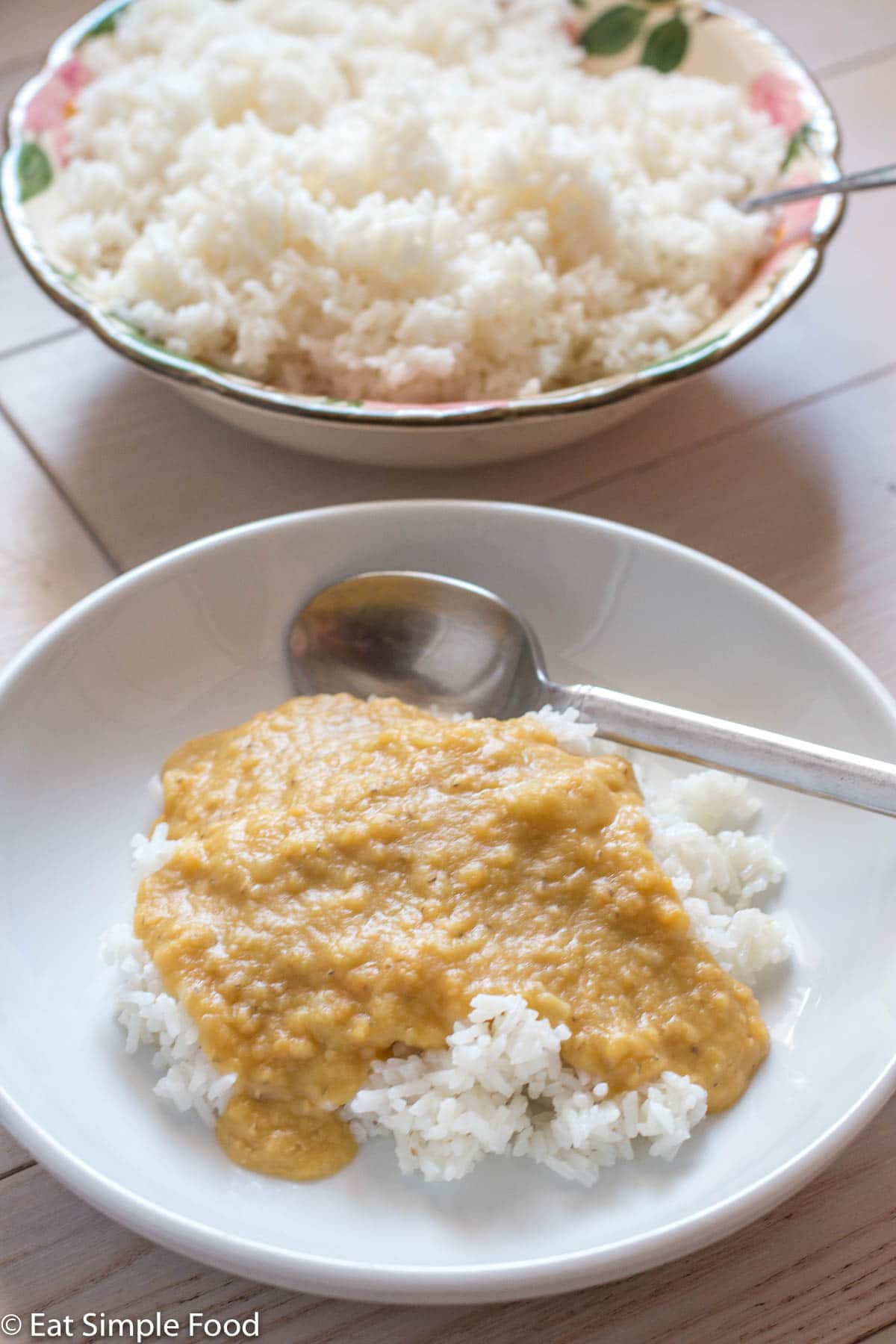 Side view. White bowl with spoon of white rice covered in yellow brown creamy lentil puree. White bowl of rice directly behind it. On white table.
