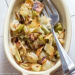 Small yellow oval bowl of chopped roasted brown fennel. Large fork in bowl. On white table. Side view. Close up.