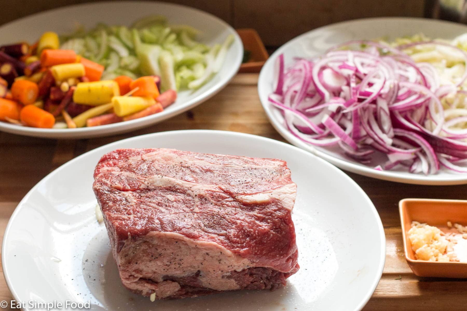 3 white plates with ingredients: raw pieces of whole chuck roast beef, multi color chopped carrots, chopped celery, sliced onions, and sliced fennel.