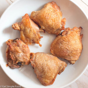 Top view of crispy browned bone in, skin on chicken thighs on a white plate on a white table.