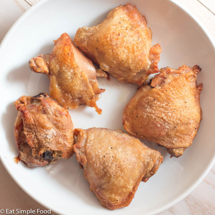 Top view of crispy browned bone in, skin on chicken thighs on a white plate on a white table.