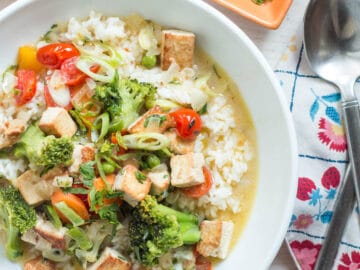 Top view of white plate/bowl of white rice covered in a yellow curry sauce with cubed tofu, broccoli, cherry tomatoes, and peas. Fork and spoon on napkin on side.