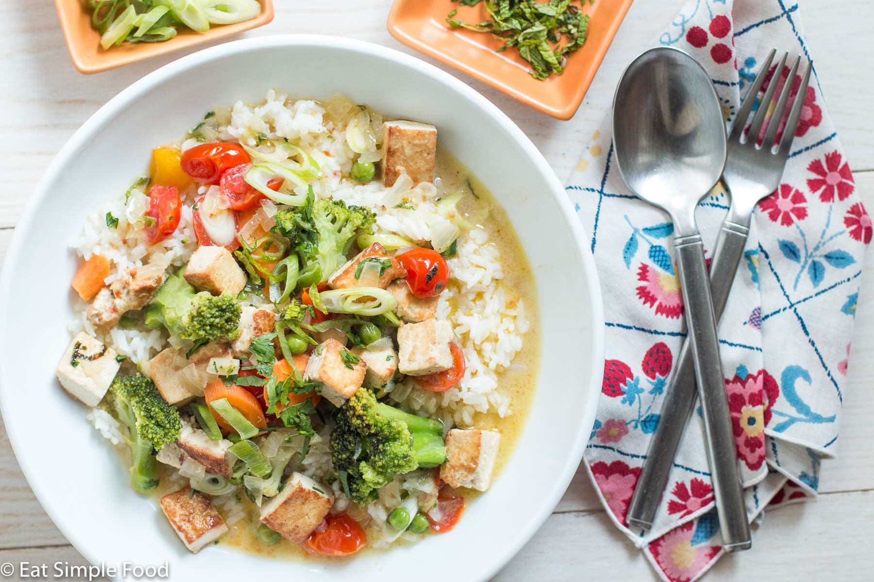 Top view of white plate/bowl of white rice covered in a yellow curry sauce with cubed tofu, broccoli, cherry tomatoes, and peas. Fork and spoon on napkin on side.