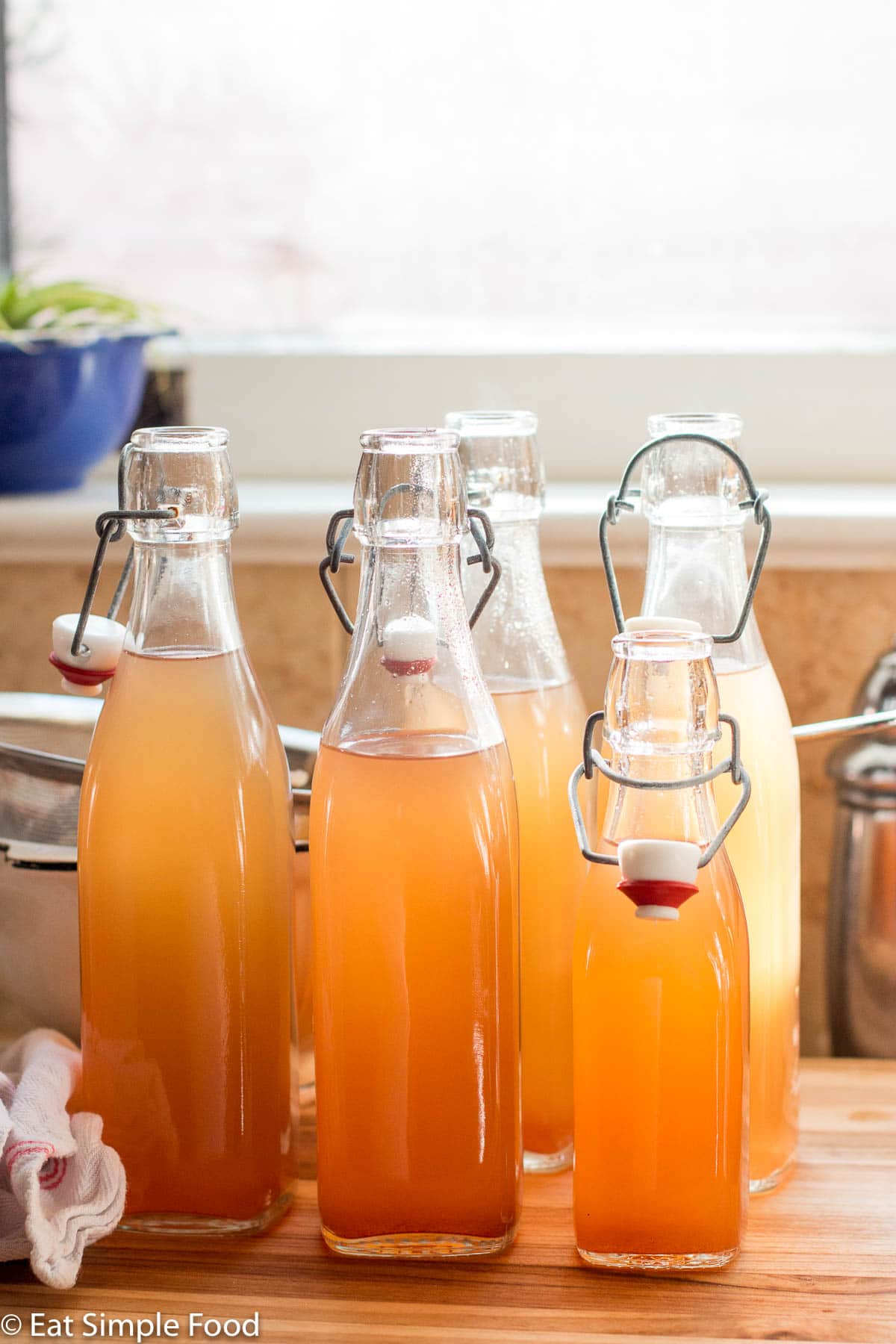 5 bottles of opened homemade apple cider on a wood cutting board in front of a window and windowsill.