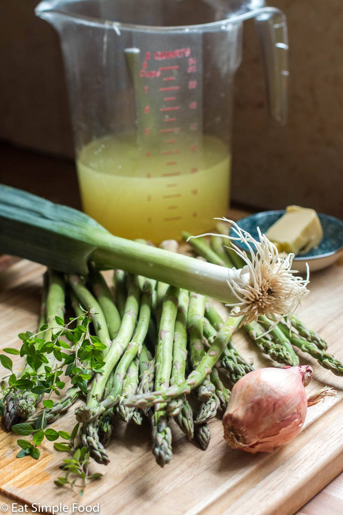 Raw Ingridients: Asparagus, leek, shallot, thyme sprigs, and a measuring cup of yellow broth on a wood cutting board.
