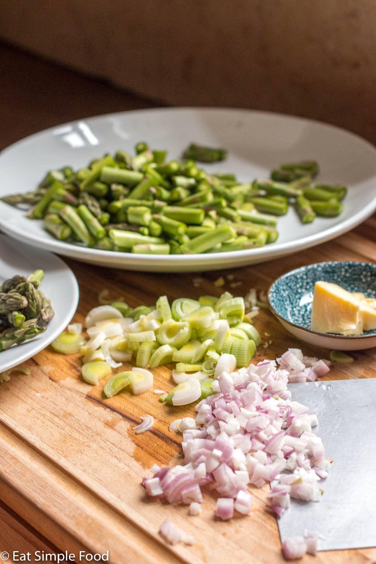 Wood chopping board with diced shallots and sliced leeks with a small container of butter and a large plate of cut up asparagus and a smaller plate of asparagus tips.