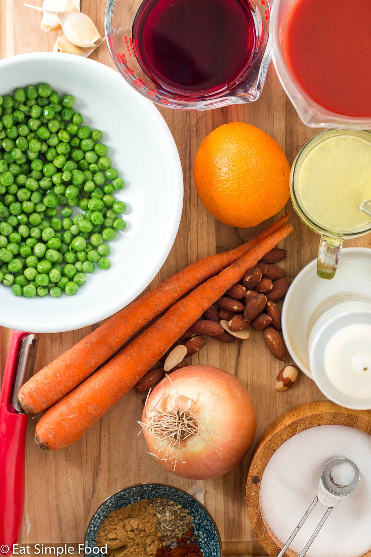 Top view of ingredients on a wood cutting board: bowl of peas, 2 carrots, whole almonds, orange, garlic cloves, spices in a small bowl, bowl of salt, cup of broth, cup of red wine, and cup of tomato juice.