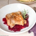 Top view of white plate with Cranberry relish on the bottom and pan fried chicken on top. Fork, knife and napkin on the side. Bowl of mashed potatoes above plate.