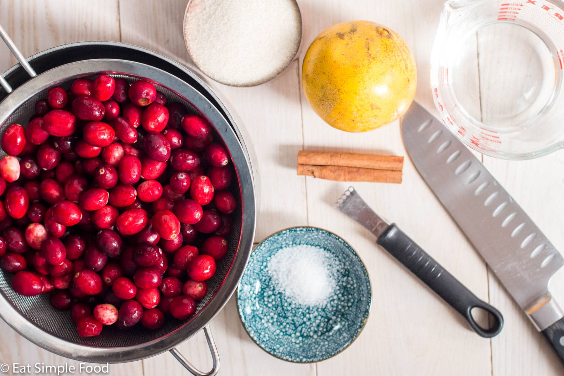 Ingredients for cranberry sauce in bowls on a white table: strainer of fresh cranberries, salt, sugar, orange, cinnamon stick, measuring cup of water, zester, and knife.