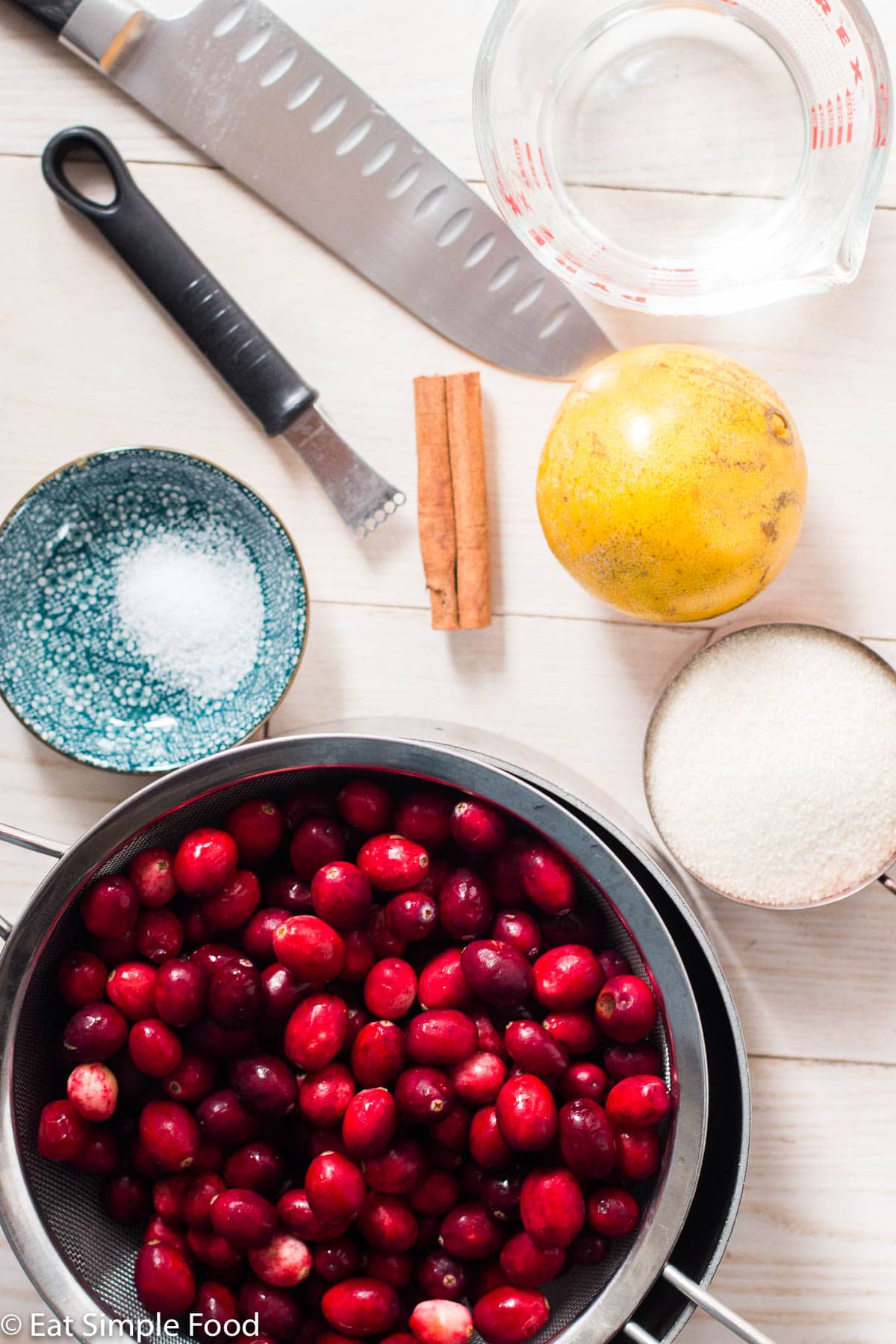 Ingredients for cranberry sauce in bowls on a white table: strainer of fresh cranberries, salt, sugar, orange, cinnamon stick, measuring cup of water, zester, and knife.