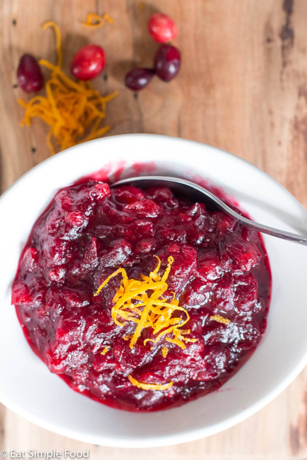 white bowl of cranberry sauce with orange zest with a spoon sticking out on a wood cutting board. Top view.