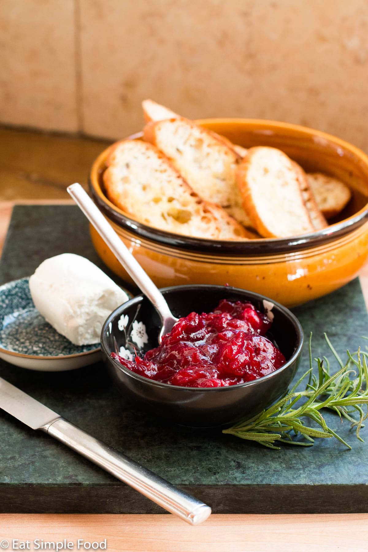 Ingredients: small bowl of cranberry sauce, bowl of toasted sliced baguette, small bowl of goat cheese log with knife. 2 sprigs of rosemary. Side view.