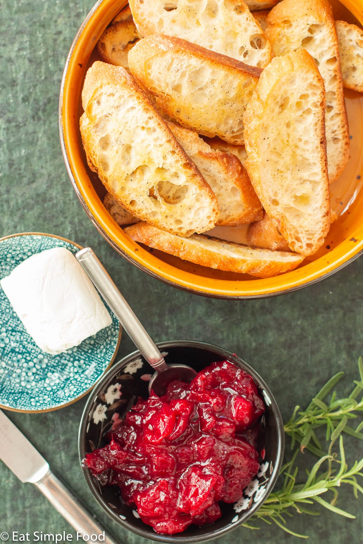 Ingredients: small bowl of cranberry sauce, bowl of toasted sliced baguette, small bowl of goat cheese log with knife. 2 sprigs of rosemary. Top view.
