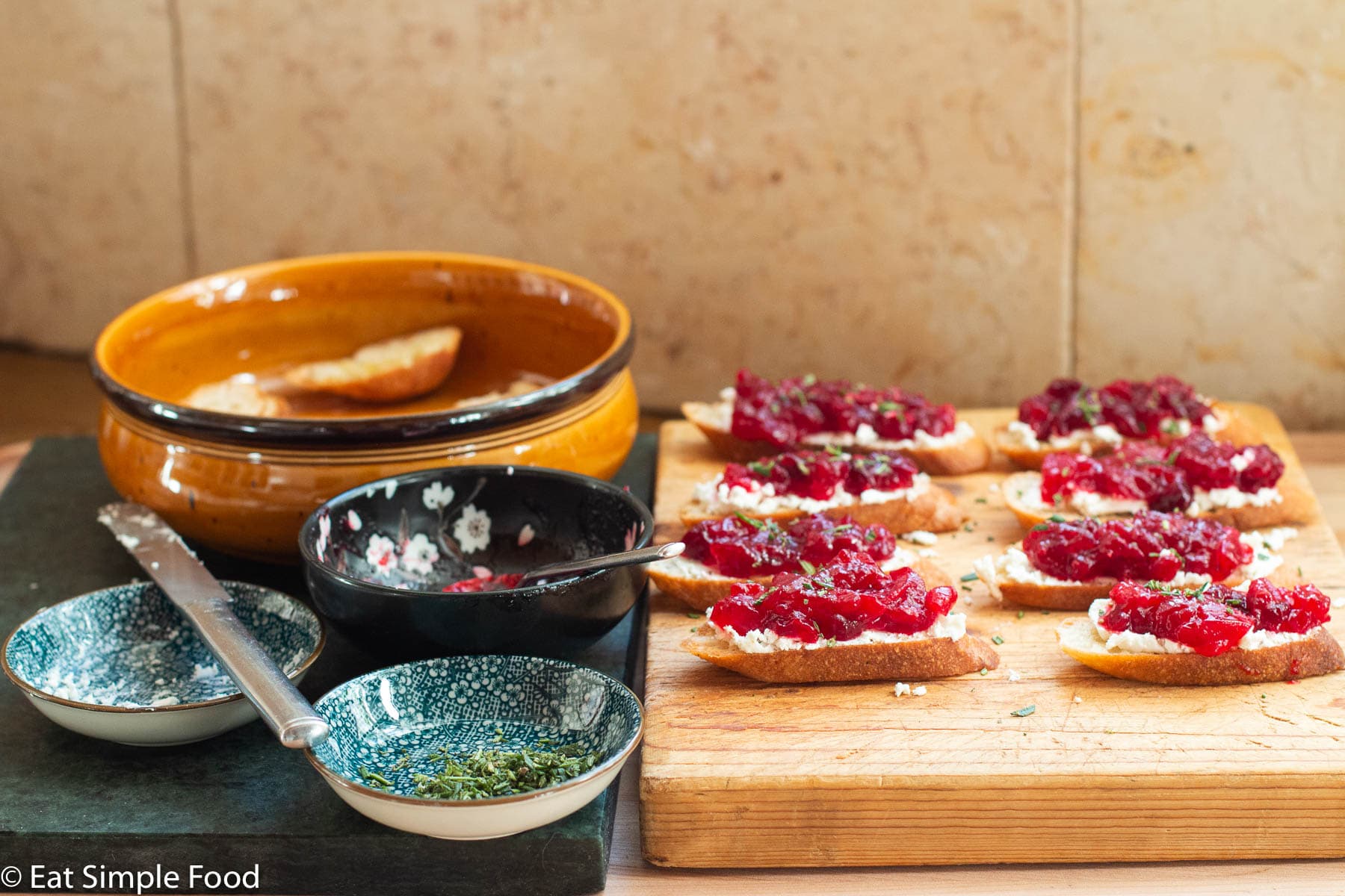 8 little crostini toast on a wood board topped with goat cheese and cranberry sauce and garnished with rosemary. Side view with bowls of ingredients on the side.