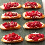 8 little crostini toast on a green slab board topped with goat cheese and cranberry sauce and garnished with rosemary. Side view.