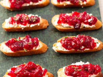 8 little crostini toast on a green slab board topped with goat cheese and cranberry sauce and garnished with rosemary. Side view.