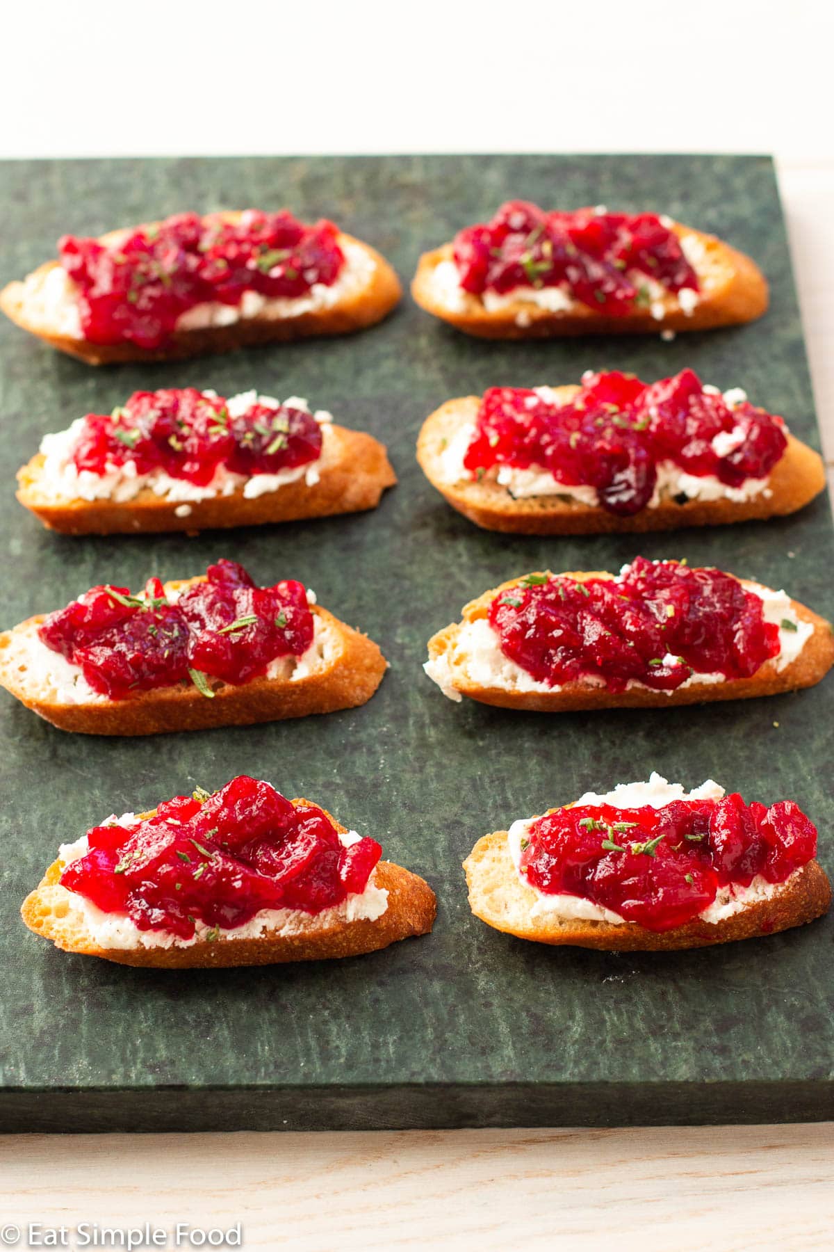 8 little crostini toast on a green slab board topped with goat cheese and cranberry sauce and garnished with rosemary. Side view.