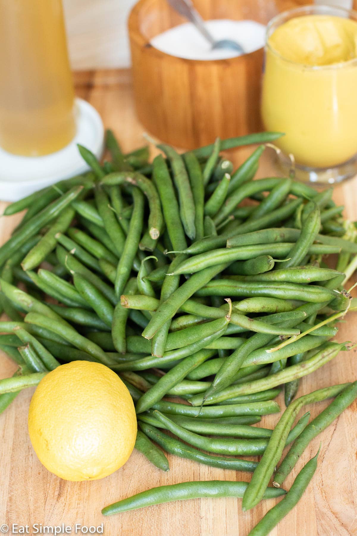 Side view of raw green beans with a lemon in front and jar of mustard, salt, and olive oil in the back. Side view.