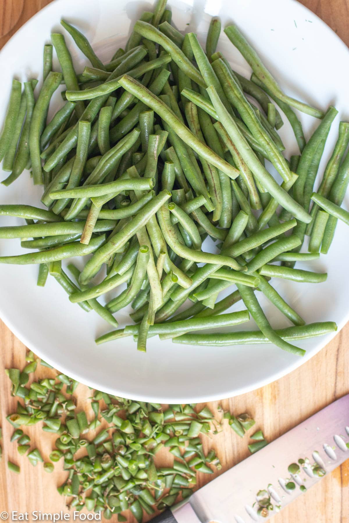 Top view of white plate of green beans with the ends cut off and laying on a wood cutting board with a chef's knife.