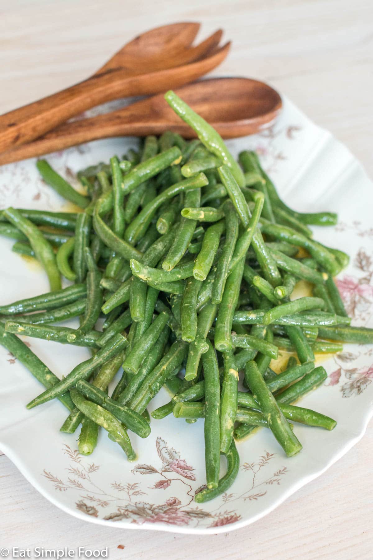 Side view of cooked green beans in a yellow sauce on a white plate with brown wood tongs on a white table.