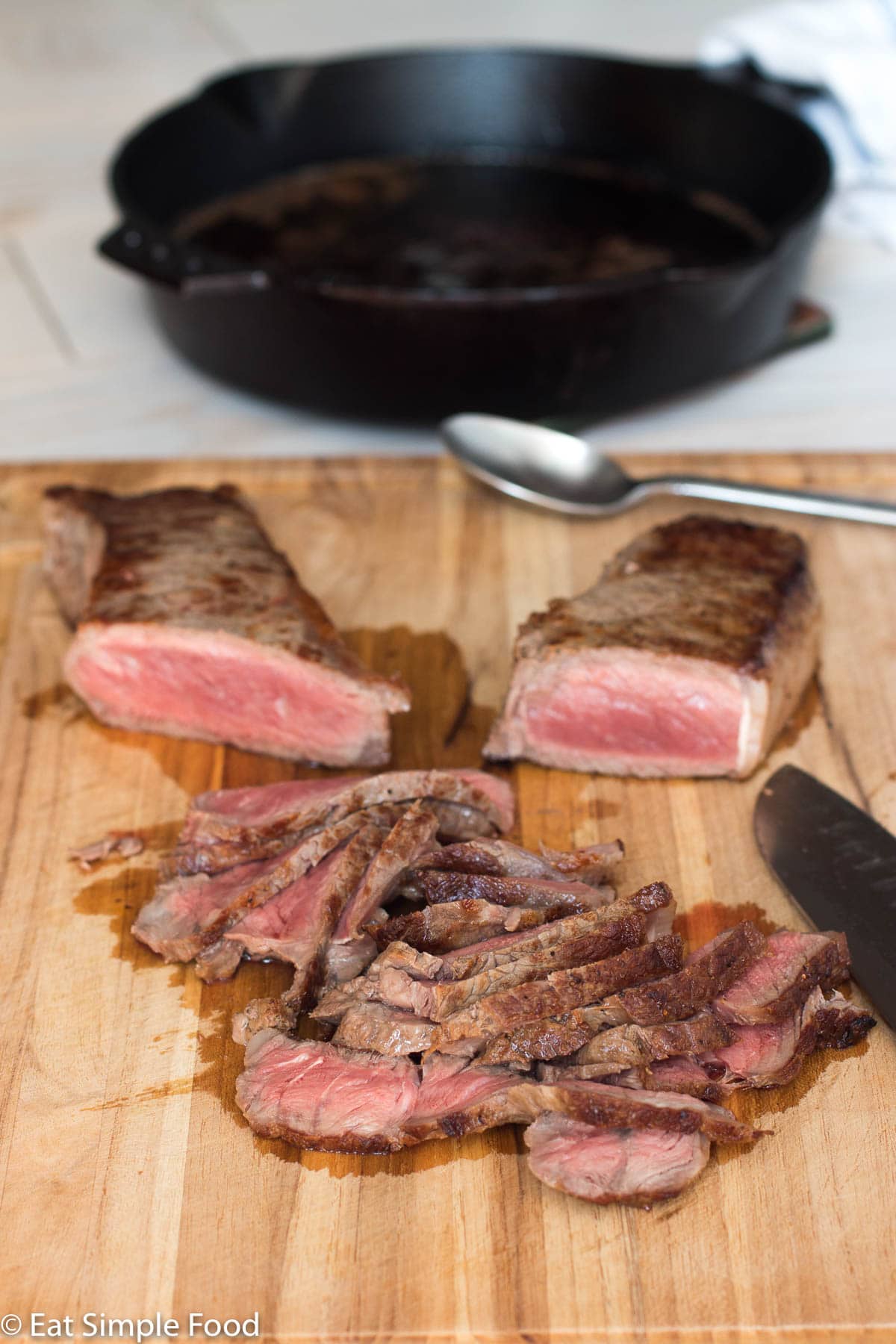 Two steaks on a wood cutting board with a chefs knife. Half of each steak is sliced and pink in the middle. Cast iron pan in background.