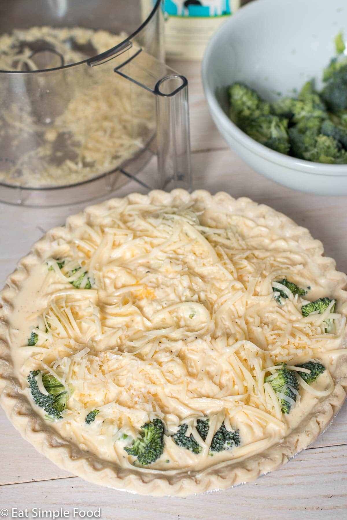 Side view of raw broccoli and swiss cheese quiche with a bowl of broccoli and a bowl of cheese and a carton of milk in the background.