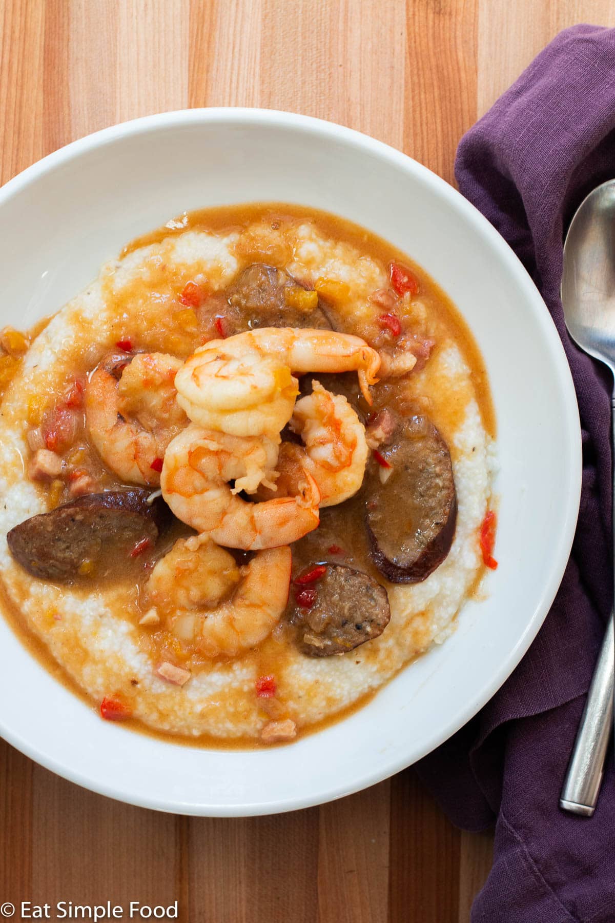 Shrimp and Andouille Sausage slices in a red brown gravy over white grits in a white bowl. Spoon and purple napkin on side.