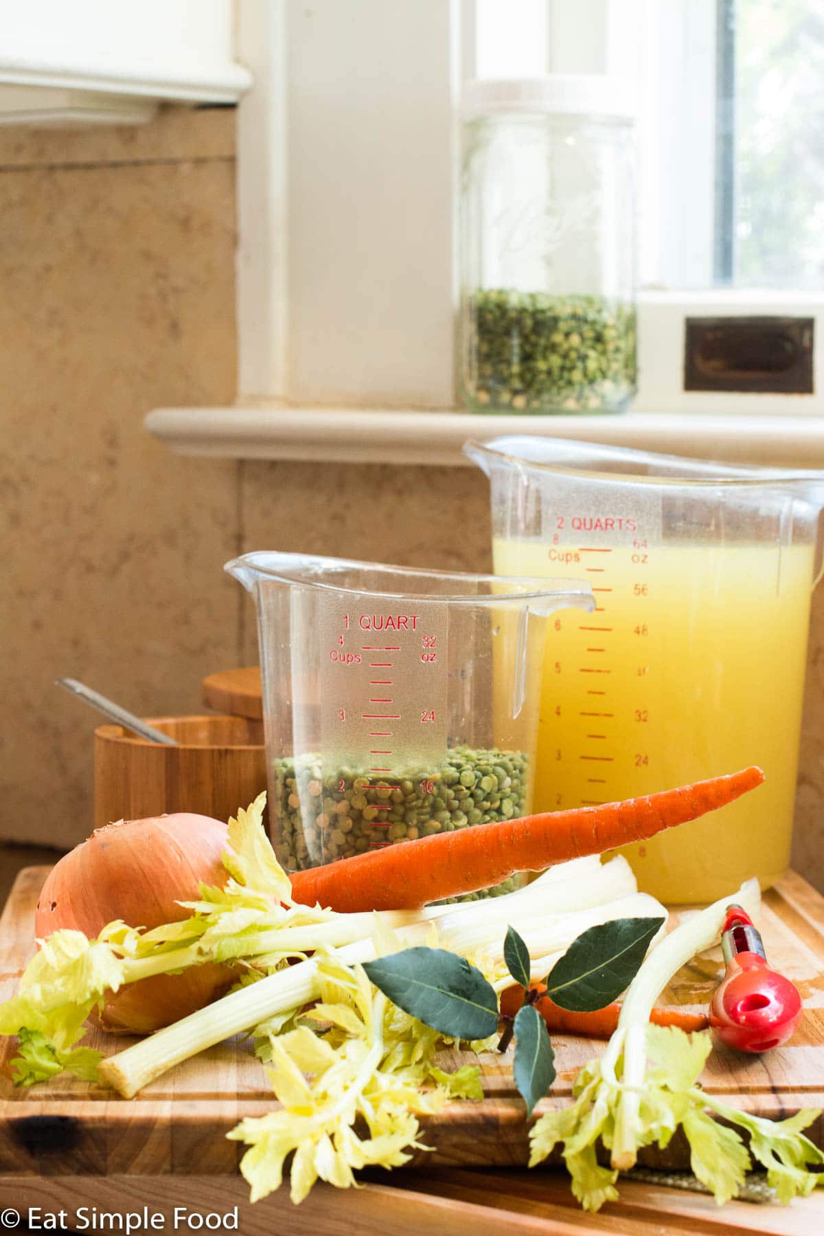 Ingredients on a wood cutting board: bay leaves, celery with leaves, onion, carrot, split peas in a measuring cup and broth in a measuring cup.