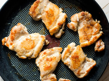 5 seared cooked boneless skinless chicken thighs in a black pan on a white table. Top down view.