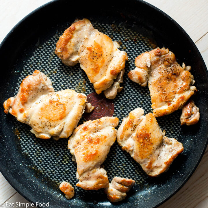 5 seared cooked boneless skinless chicken thighs in a black pan on a white table. Top down view.