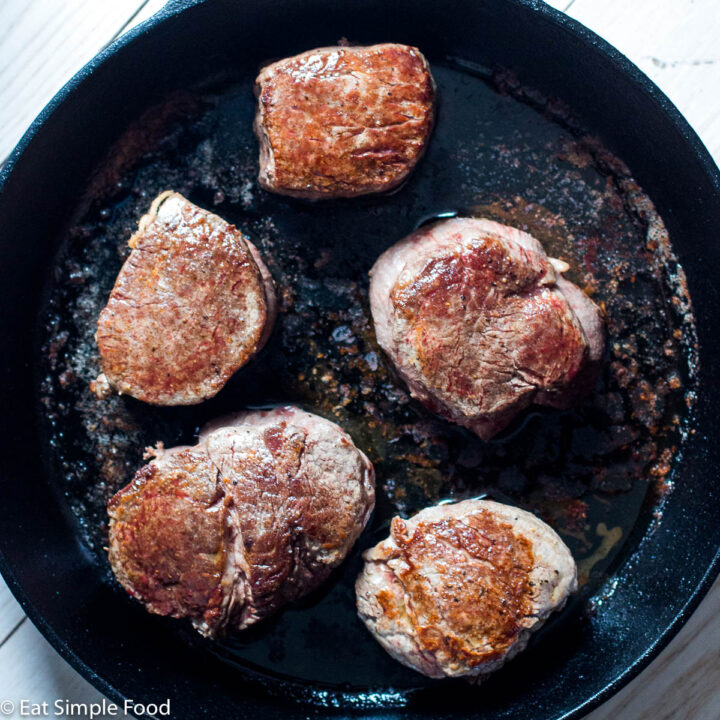 Top view of five pieces of cooked seared filet mignon in a cast iron pan on a white table